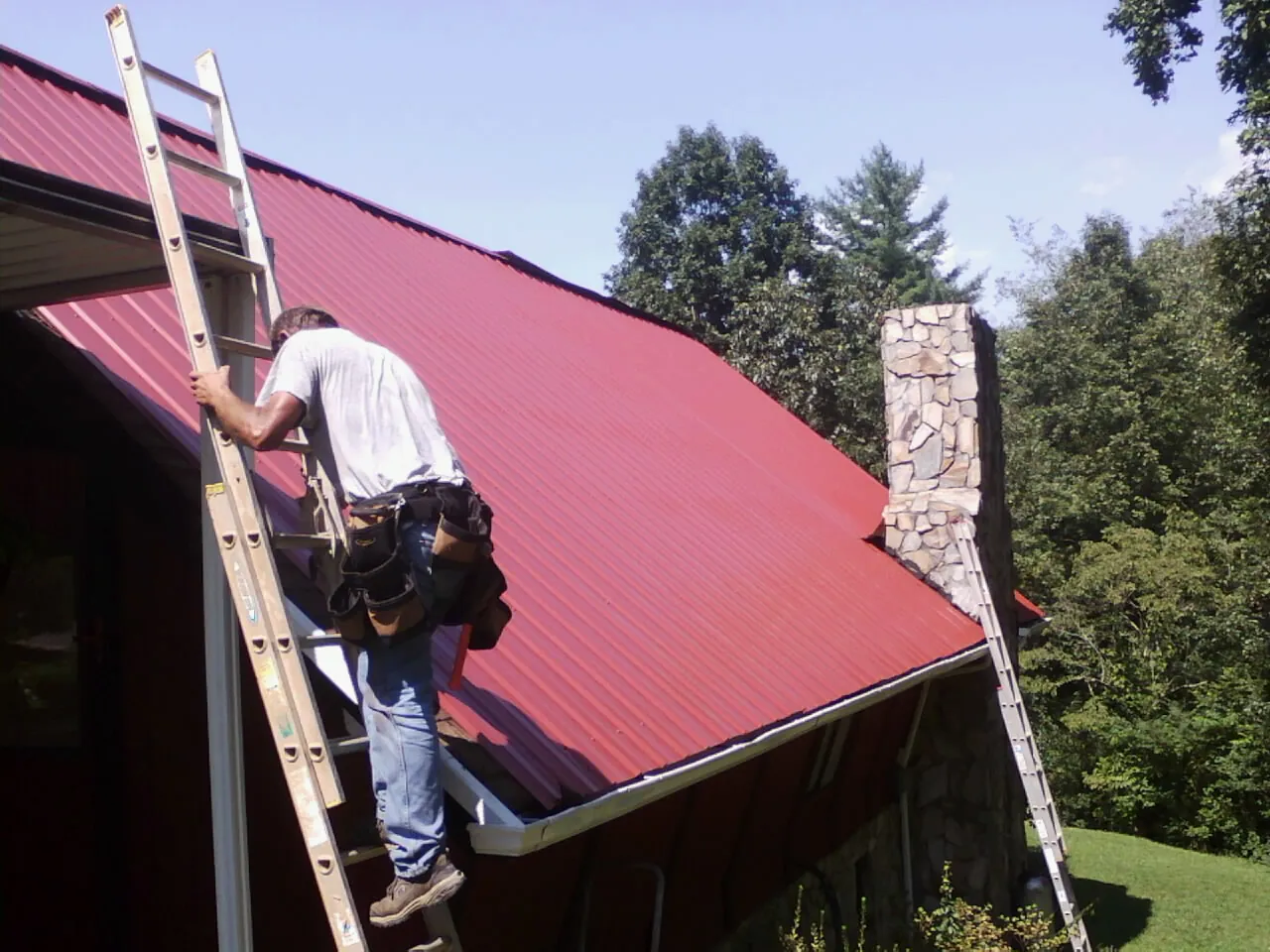 Roofer on a ladder beside a red metal roof and stone chimney — Soffit & Fascia Repair by Roofing Brothers in Mill Hall